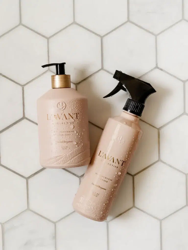 Two bottles of L'AVANT's High Performing Hand Soap in Blushed Bergamot are placed on a white hexagonal tile background. One bottle has a pump dispenser, while the other features a spray nozzle. Both bottles are made of pinkish-beige plastic and appear slightly wet with water droplets.