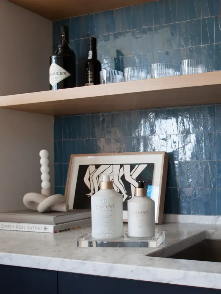 A stylish bathroom countertop with Lavant lotion and soap bottles elegantly arranged on a medium Lucite Tray by L'AVANT Collective on a marble surface. Above the counter, wooden shelves hold various decor items, including a framed abstract print, glassware, and bottles. The backsplash is made of blue tile.