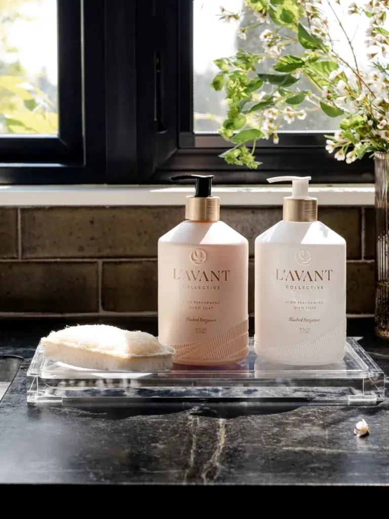 Two bottles of High Performing Hand Soap - Blushed Bergamot from L'AVANT Collective are placed on a clear tray on a dark countertop. On the left is a peach-colored bottle with a pump, and on the right is a white bottle with a pump. A round sponge sits on the tray beside the bottles, and a plant is visible in the background.