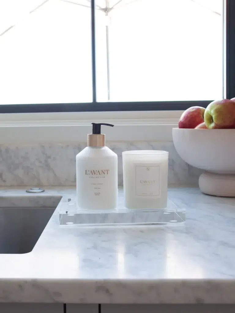 A marble counter beside a stainless steel sink features a Lucite Tray - Medium by L'AVANT Collective, elegantly holding hand soap and a candle in white containers with gold accents. In the background, a bowl filled with apples sits on the counter near a window.
