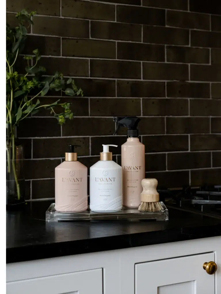 A stylish kitchen countertop with a dark tile backsplash features a tray showcasing L'AVANT Collective cleaning products: a High Performing Hand Soap in Blushed Bergamot scent, a dish soap, a multi-purpose cleaner, and a wooden brush. Adjacent greenery adds a touch of nature.