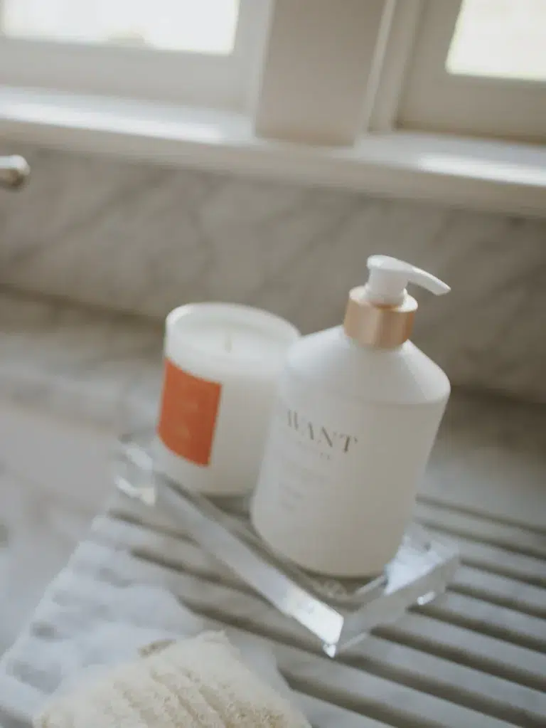 A Lucite tray from L'AVANT Collective on a countertop next to a window holds a white soap dispenser with an elegant pump and a candle in a white jar with an orange label. In the foreground, there is a folded towel.