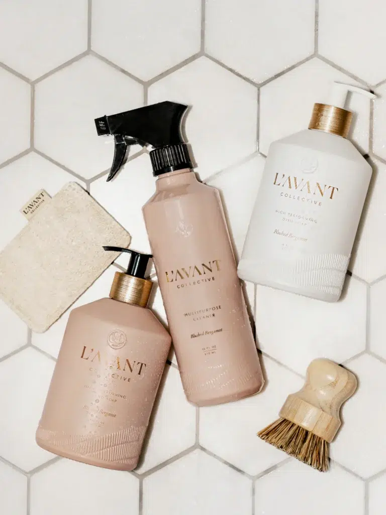 Three bottles of L'AVANT Collective cleaning products are displayed on a white hexagonal tiled surface. The bottles include a spray cleaner, the High Performing Hand Soap - Blushed Bergamot in a pump bottle, and a bottle with a screw top. A scrubbing brush and a natural loofah cloth are also arranged around the bottles.