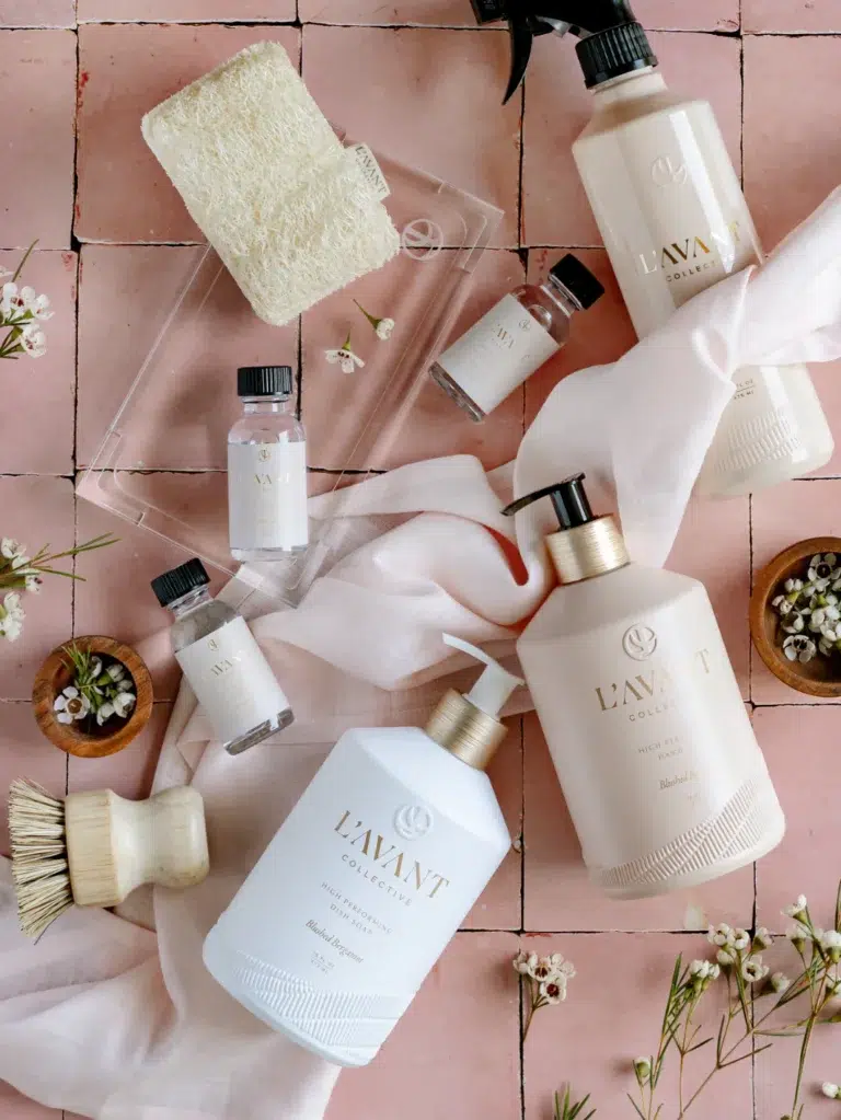An arrangement of L'AVANT Collective skincare and beauty products, featuring the High Performing Hand Soap in Blushed Bergamot, bottles of various sizes, a bar of soap, a small brush, and a clear tray. The items are displayed on a pink tiled surface with sprigs of small white flowers and a white cloth.