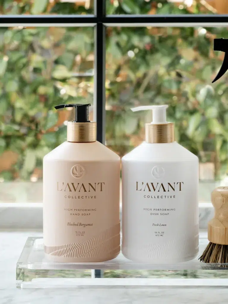 Two bottles of L'AVANT Collective soap are placed on a marble counter in front of a window with greenery outside. One bottle is labeled "High Performing Hand Soap - Blushed Bergamot - L'AVANT Collective," and the other is labeled "High Performing Dish Soap, Fresh Lime.