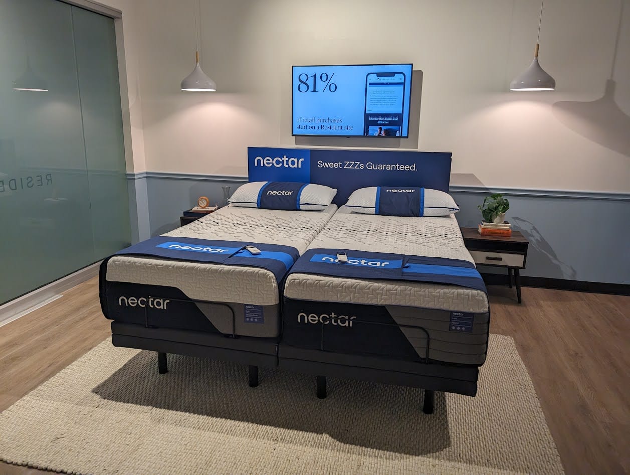 A showroom display with two Nectar mattresses set up on adjustable bases. The mattresses are side by side, each covered with white and blue bedding. A large sign above the beds reads, "Sweet ZZZ's Guaranteed." A nightstand with decorations is situated beside the beds, perfect for your next Nectar Mattress Review.