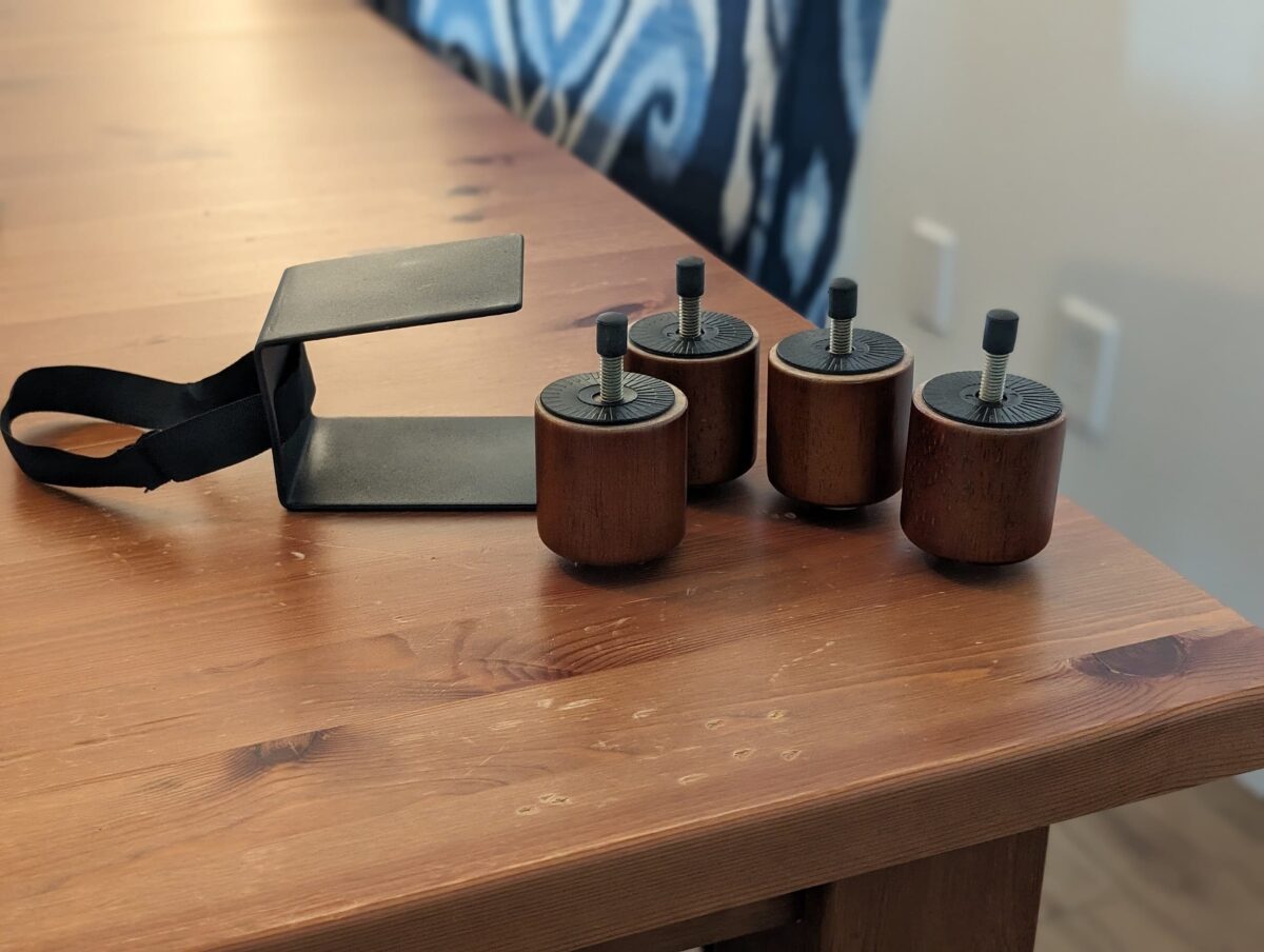 Four wooden furniture legs with screw-on attachments are placed on a wooden table. Next to them, a black metal bracket with a strap lays flat on the surface. The background is slightly blurry, revealing part of a chair with patterned fabric—a perfect scene for any homebody’s cozy couch setup.