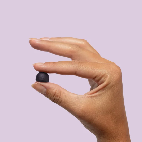 A hand with neatly trimmed nails is holding a small, black, rounded WonderSleep Mushroom Gummy - Plant People between the thumb and forefinger against a plain, light purple background.