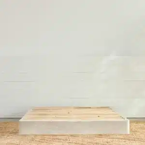 A minimalist bedroom setting featuring the Avocado Foundation, a low wooden bed frame without a mattress. The bed frame is placed on a natural woven rug, with white shiplap paneling on the wall behind, and soft light casting gentle shadows across the setting.