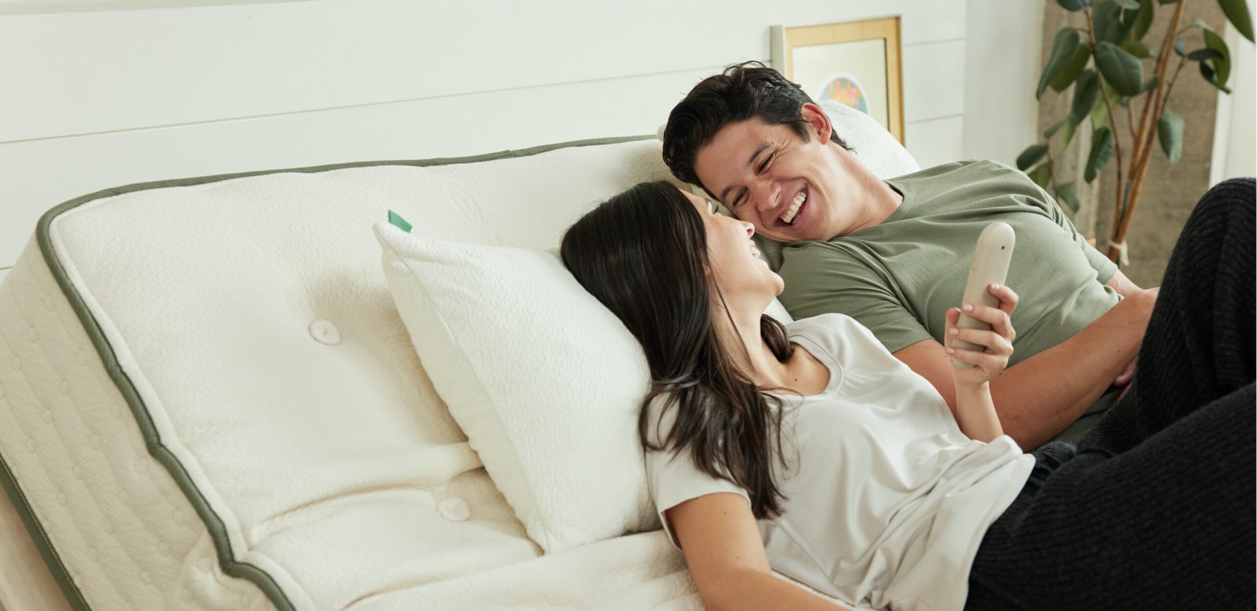 A couple is lying on a sofa bed, looking at each other and smiling. The woman is holding a remote control that appears to be for adjusting the bed. They are in a cozy, well-lit room with a plant and framed artwork in the background.