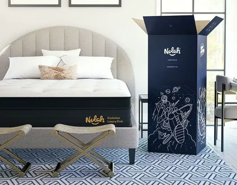 A bedroom with a bed featuring a beige upholstered headboard and a Nolah mattress on a black base labeled "Nolah Evolution Luxury Firm." The bed is flanked by stools and set on a geometric-patterned rug. A Nolah-branded box stands nearby, partially opened.