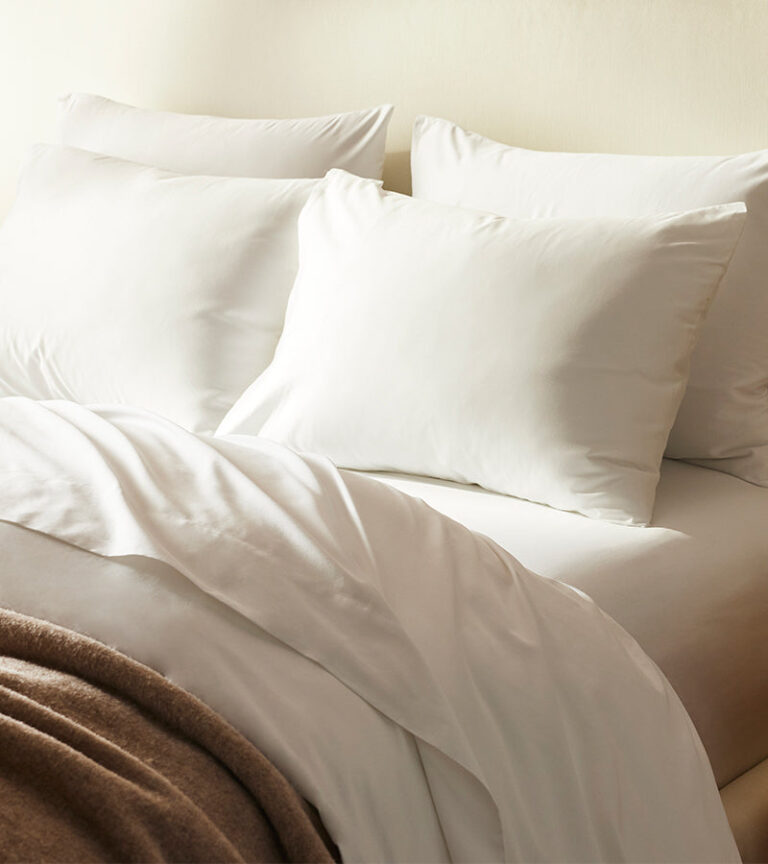 A neatly made bed with white pillows and a white duvet. A brown blanket is folded over the edge of the bed. The bed is positioned against a light beige wall, creating a minimalist and cozy atmosphere.