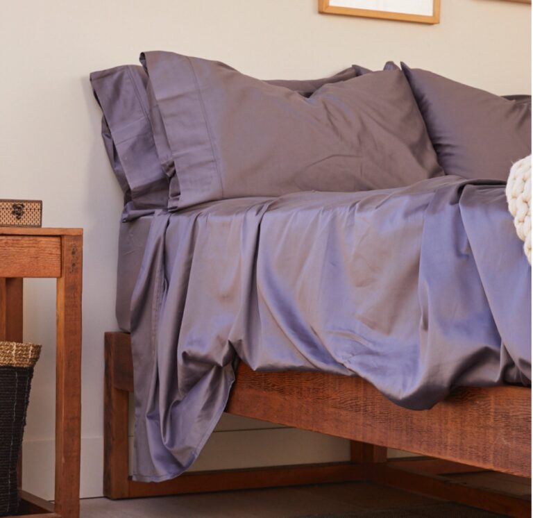 A neatly made bed with grey sheets and pillowcases, slightly wrinkled, is shown next to a wooden nightstand with a woven basket and small decorative items on top. The bedspread hangs over the edge of the wooden bed frame. A soft, cream-colored blanket is draped on the far side.