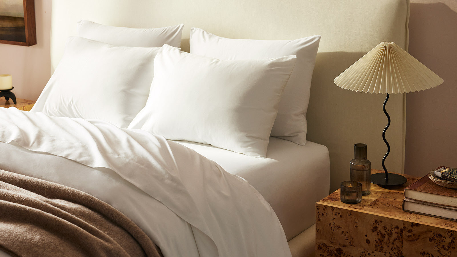 A neatly made bed with white sheets and pillows is adjacent to a wooden nightstand holding a pleated lampshade lamp, a glass bottle, and stacked books. A cozy beige blanket is draped over the bed, creating a warm and inviting bedroom atmosphere.