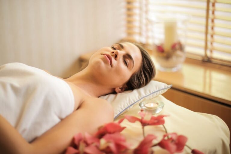 A woman with her eyes closed is lying on a spa bed. She is wrapped in a white towel and appears relaxed. In the foreground, there are pink flowers, while in the background, a large candle and a window with blinds can be seen.