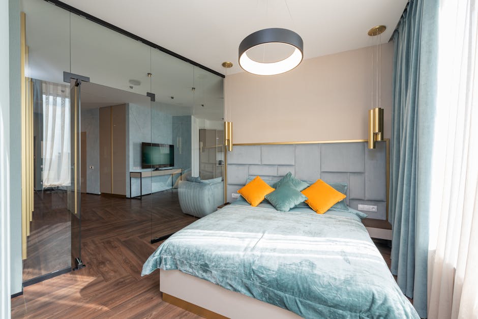 Modern bedroom featuring a bed with teal bedding and two orange throw pillows. The room includes a large, circular ceiling light, floor-to-ceiling windows with teal curtains, and a glass divider separating a living area with a television and cabinetry.