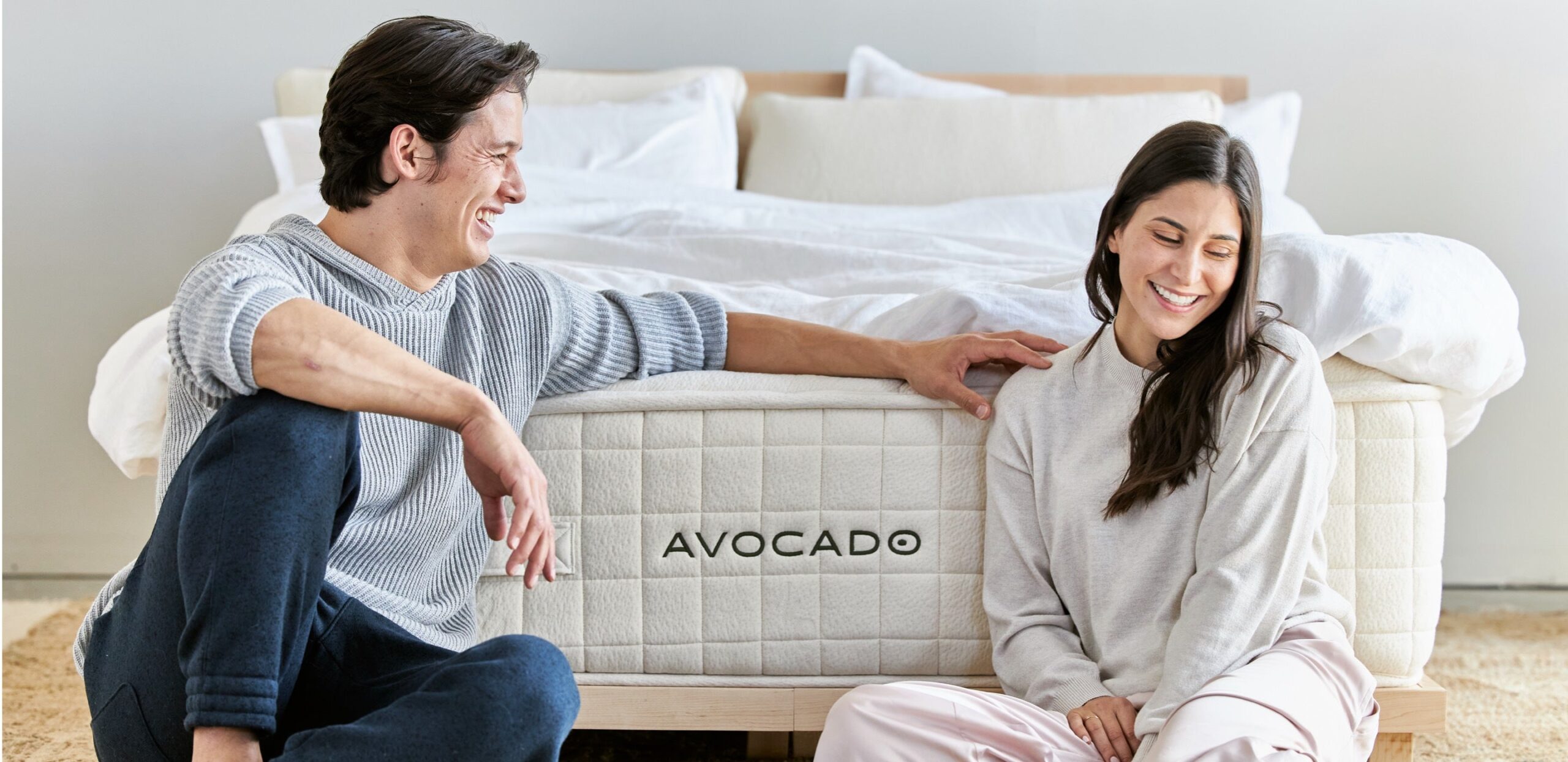 A smiling man and woman sit on the floor next to each other, leaning on the base of a bed. The bed has a white quilted mattress with the word "AVOCADO" written on it. The man is wearing a gray sweater and the woman is in a light-colored long sleeve top and pants.