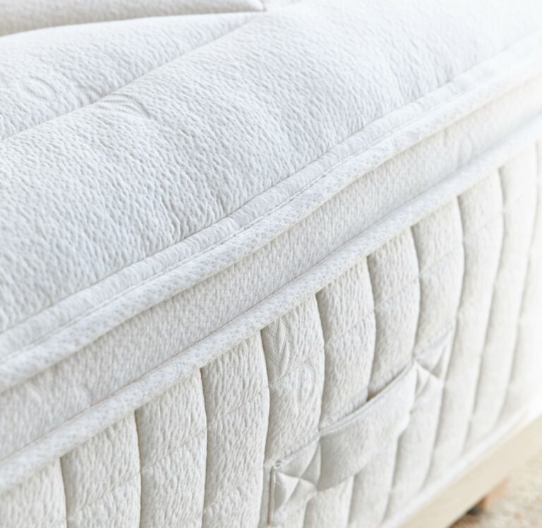 Close-up image of a white, quilted mattress featuring a plush pillow-top layer. The mattress has a textured fabric design with detailed stitching and a side pocket for convenient storage. The image highlights the mattress's comfortable and high-quality build.