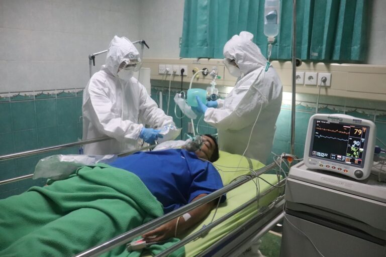 A patient wearing an oxygen mask lies on a hospital bed, covered with a green blanket. Two healthcare workers in full protective gear attend to the patient. Medical equipment, including a heart rate monitor, is visible nearby. The room has green curtains and walls.