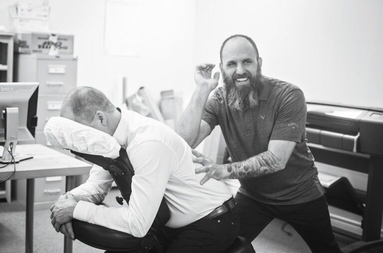 A bearded man gives a seated massage to another man who is bent forward on a massage chair with his face resting in the head support. They are in an office setting with desks, a computer, and filing cabinets in the background.