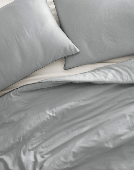 A bed is dressed with light gray satin sheets and two matching pillows. The texture of the satin material is smooth and shiny, reflecting light softly. The duvet is slightly folded back, revealing a portion of the light-colored fitted sheet underneath.