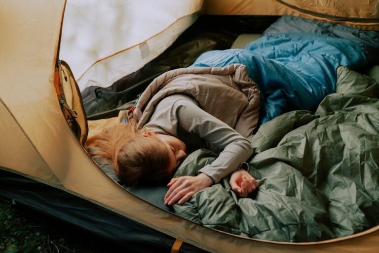 A person with long hair is sleeping inside a tent, wrapped in a blue sleeping bag, with their head resting on a green sleeping pad. The tent is partially open, revealing the outdoor environment. The person appears peaceful and comfortable.