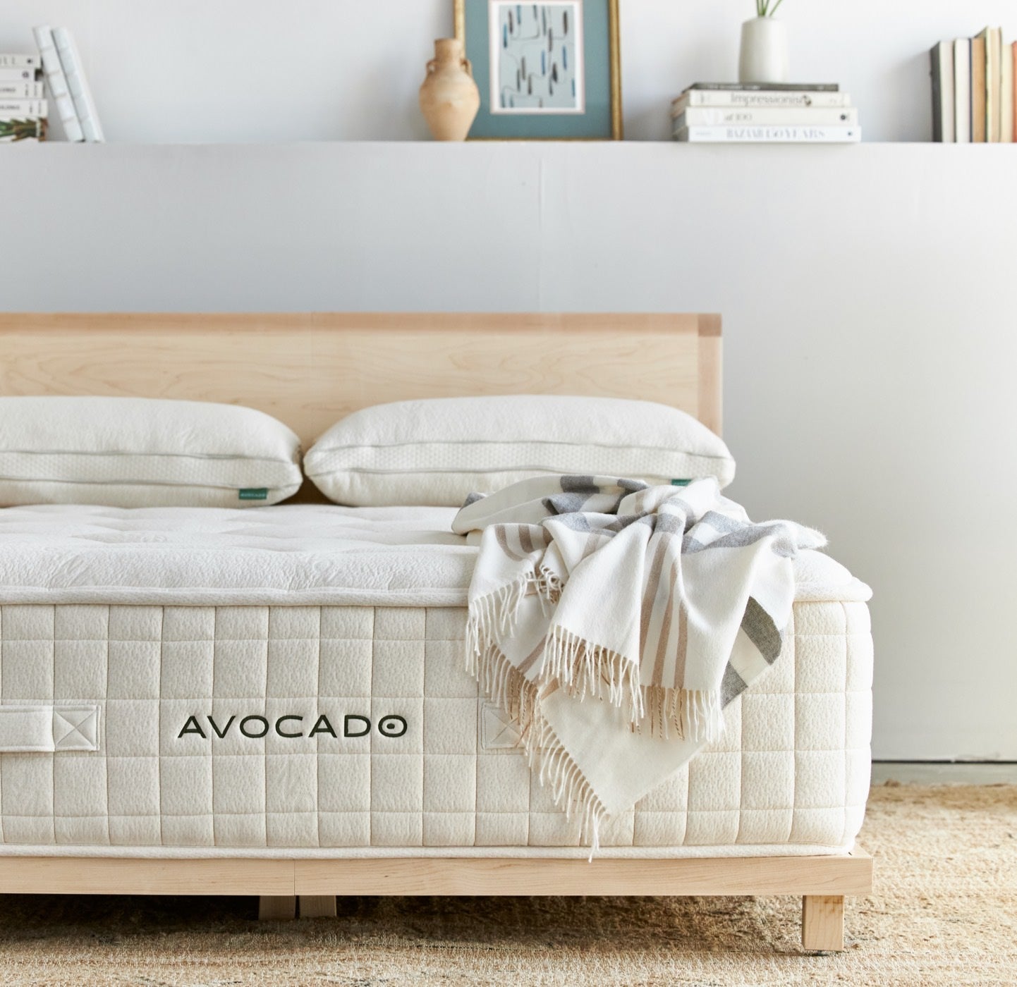 A neatly made bed with a wooden frame and a mattress labeled "AVOCADO." Two pillows are aligned at the head, and a fringed blanket is draped over the foot of the bed. The wall behind features a few books, a vase, and framed art pieces.