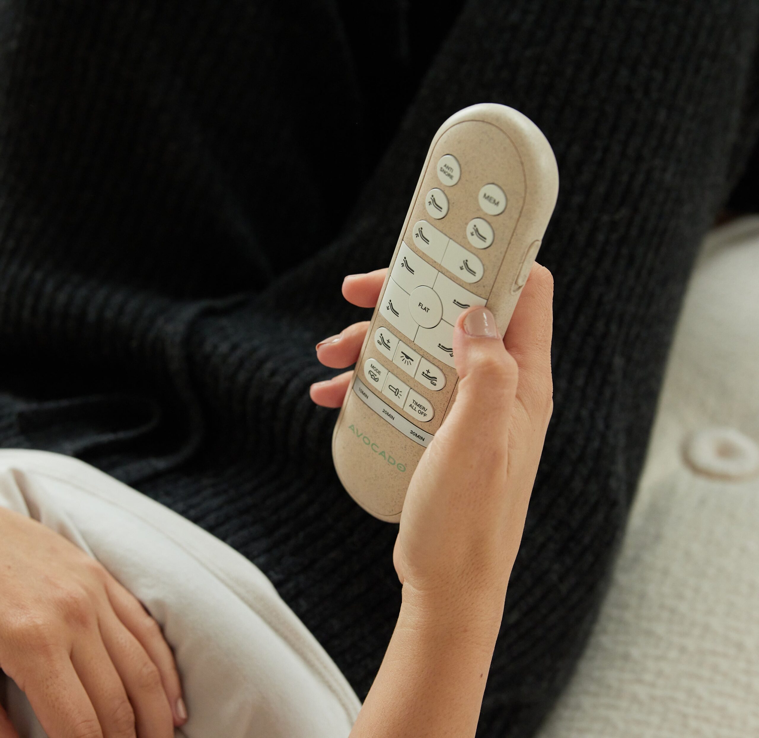 A person reclines on a couch or bed, wearing dark clothing, and holds a remote control in their right hand. The remote has several buttons labeled with functions like "Sleep" and "Auto." Only the lower half of the person's torso and hand are visible.