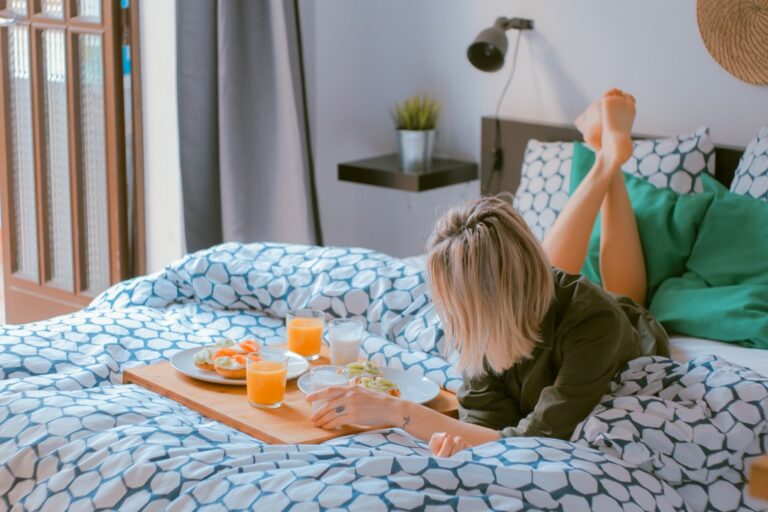 A person with blonde hair lies on a bed covered with geometric-patterned blue and white bedding. They are propped up on their elbows, reading or looking at something. On the bed in front of them is a breakfast tray with juice, food, and a plate.