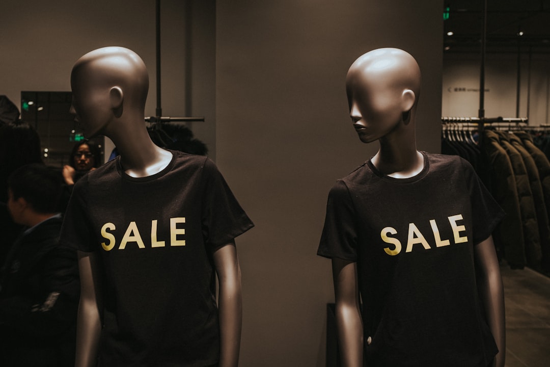 Two mannequins in a dimly lit clothing store are dressed in black t-shirts with the word "SALE" printed in bold yellow letters, indicating a sales event. Additional clothing items are visible in the background.