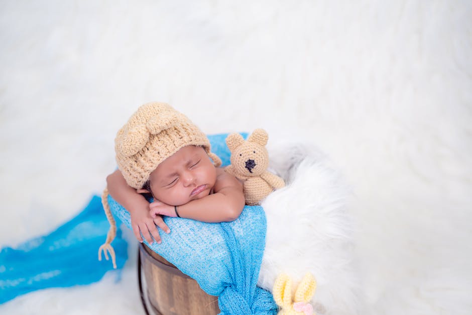 A sleeping baby, wearing a knitted bear hat, rests comfortably in a wooden bucket lined with fluffy white material. The baby is swaddled in a blue blanket, with a small knitted teddy bear tucked close by. The background is soft and white, adding to the cozy atmosphere.