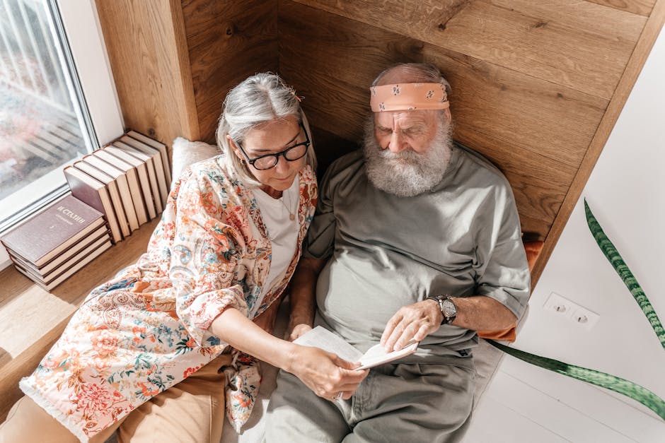 A senior woman with glasses and a floral shirt sits beside an elderly man with a beard and headband. They are sitting on a window seat with books stacked nearby, reading a book together. The cozy setting also includes a potted plant.