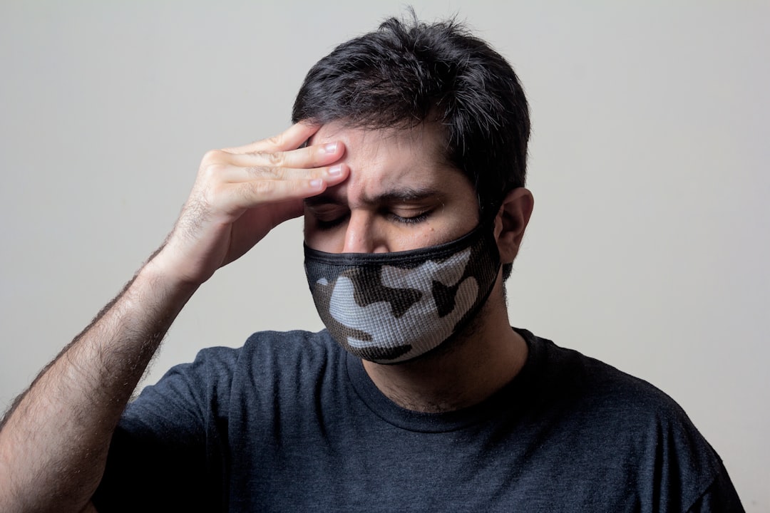 A person with short dark hair, wearing a black and white camouflage face mask and a dark shirt, is holding their forehead with a pained expression, as if experiencing discomfort or a headache. They are set against a plain light background.