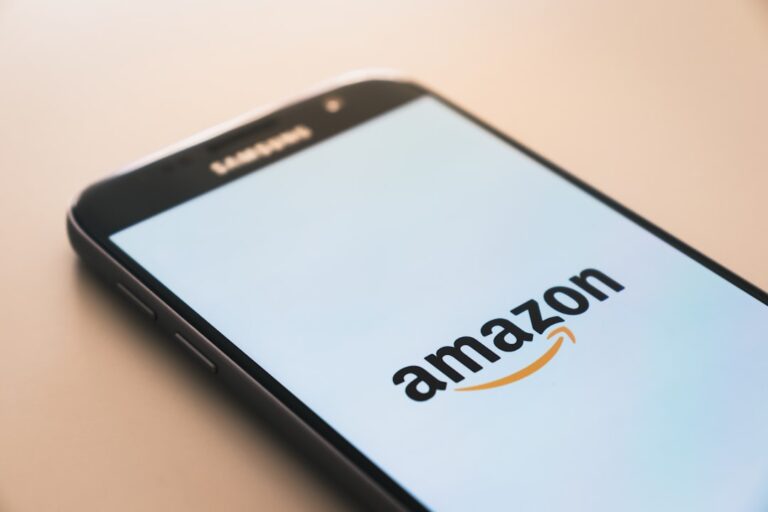 A smartphone displaying the Amazon logo on its screen lies on a light-colored surface. The phone is black and positioned at a slight angle, emphasizing the logo prominently in the center of the screen.