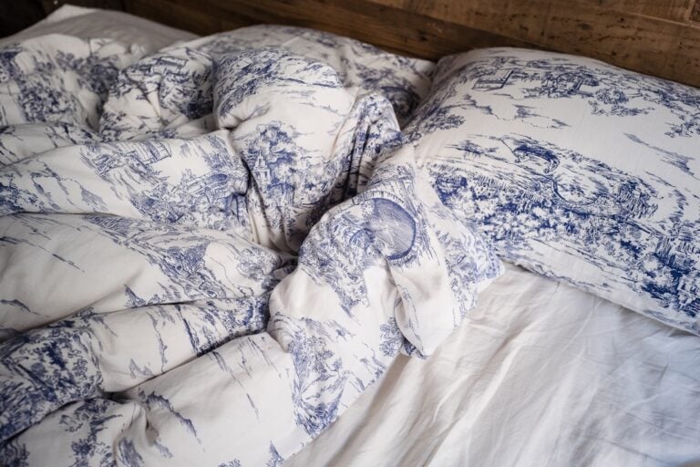 A close-up of a bed with white and blue patterned bedding. The comforter and pillowcases feature intricate design work, displaying scenes with landscapes and buildings. The headboard is wooden, and only part of the white mattress is visible.