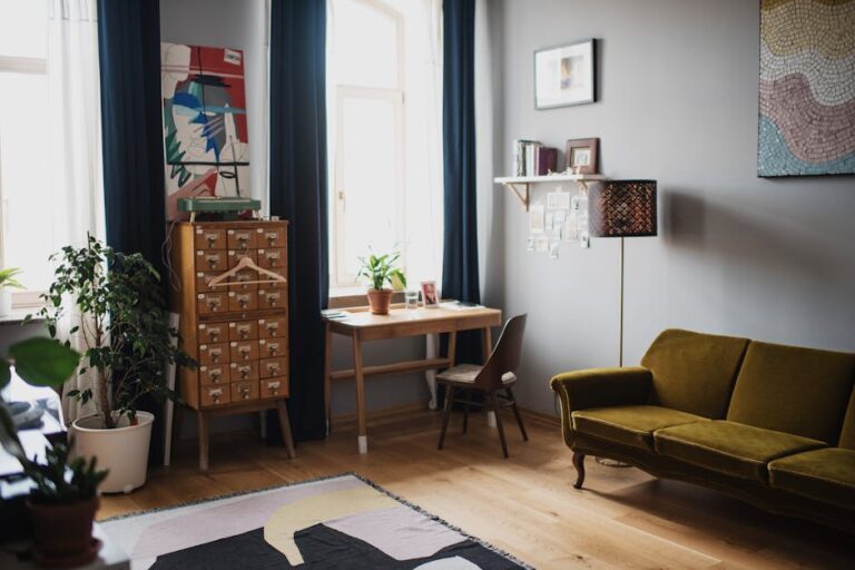 A cozy living room with a wooden floor features a green velvet couch, a small wooden desk with a plant and various items, a tall cabinet with multiple drawers, and an abstract rug. Large windows with dark blue curtains let in natural light, brightening the room.