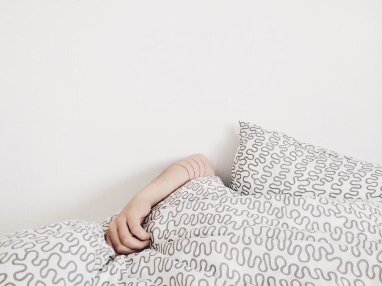 A person is partially covered in a bed with white and gray patterned bedding. Only an arm is visible, resting on the edge of the duvet. The background is a plain white wall.