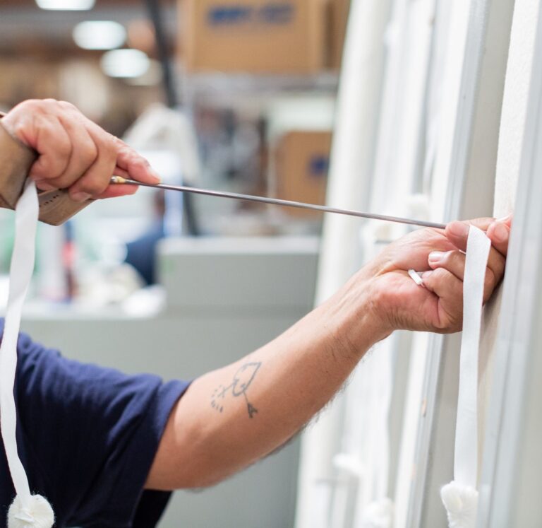 Close-up of a person threading a needle through white fabric. The individual's arm has a tattoo of a heart with wings, and they are wearing a dark blue shirt and a ring. The background features blurry industrial equipment and cardboard boxes.