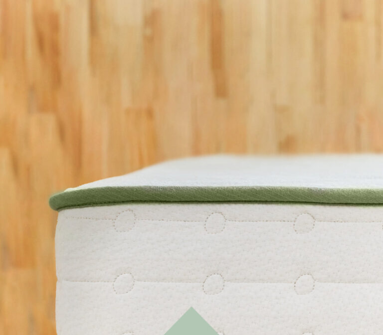 Close-up view of the corner of a mattress with a white quilted surface and a light green edge. The background is a wooden floor with a blurred effect.