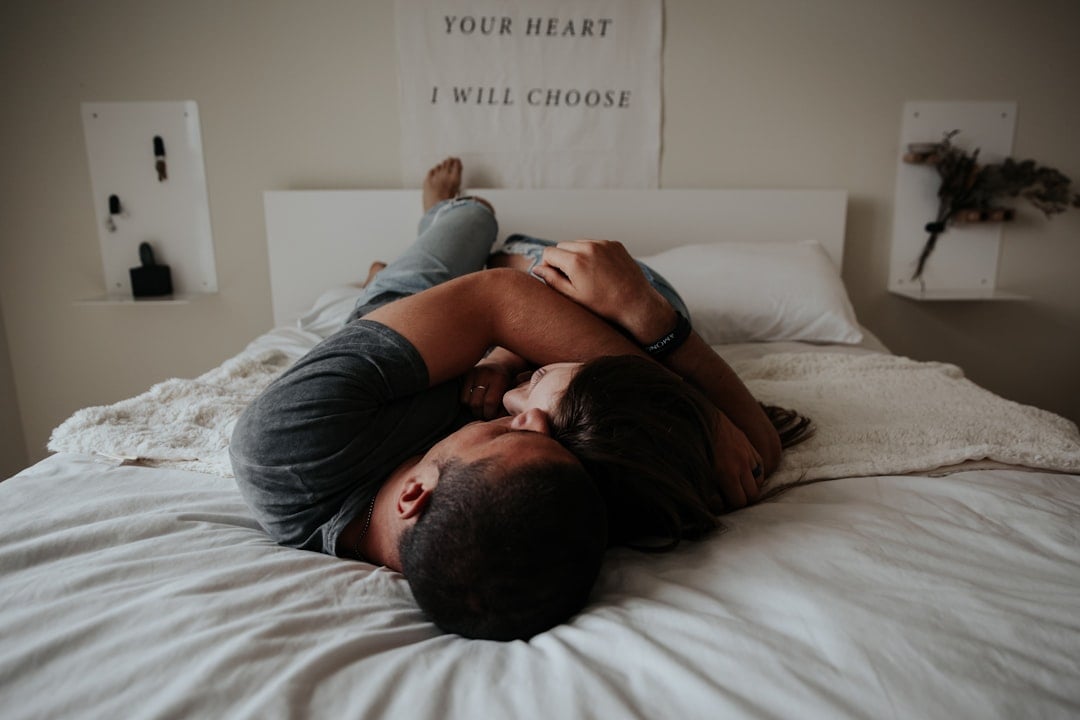 A couple lies on a bed, closely embracing. They are covered by a blanket with various pillows around them. Above the headboard, a wall hanging displays the phrase "YOUR HEART I WILL CHOOSE." The ambiance suggests intimacy and comfort.