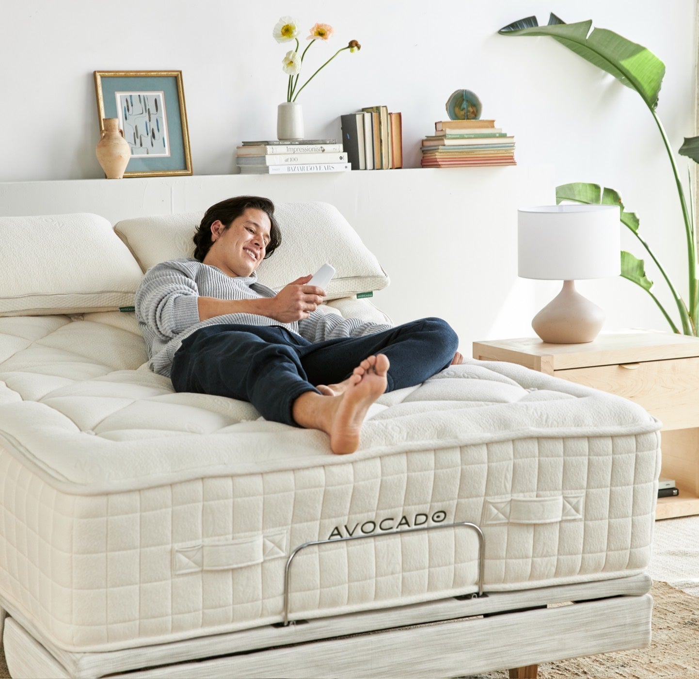 A person is lying on a bed with a white mattress, which has "AVOCADO" written on the side. The individual is holding a phone and smiling. The bedroom has a modern aesthetic with books, plants, a bedside table, and a lamp.