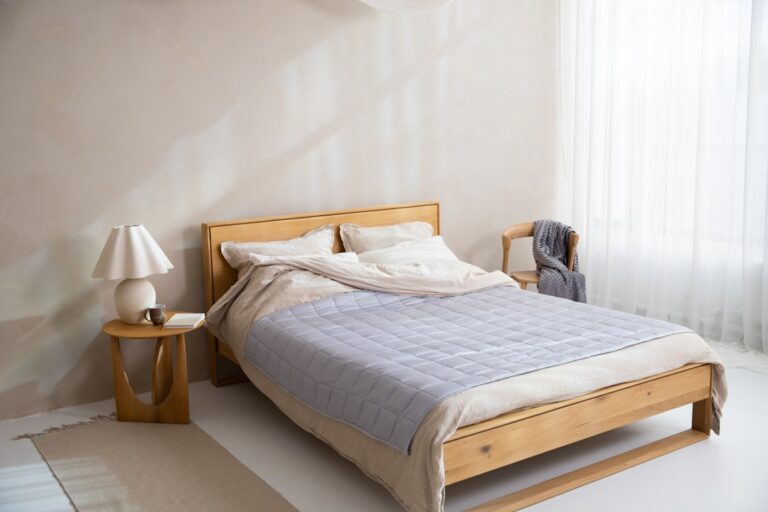 A minimalist bedroom with a light wooden bed frame, beige bedding, and a light gray quilt. A wooden bedside table holds a white lamp and a book. The room is well-lit by natural light through sheer white curtains. A cozy chair with a blanket is in the corner.