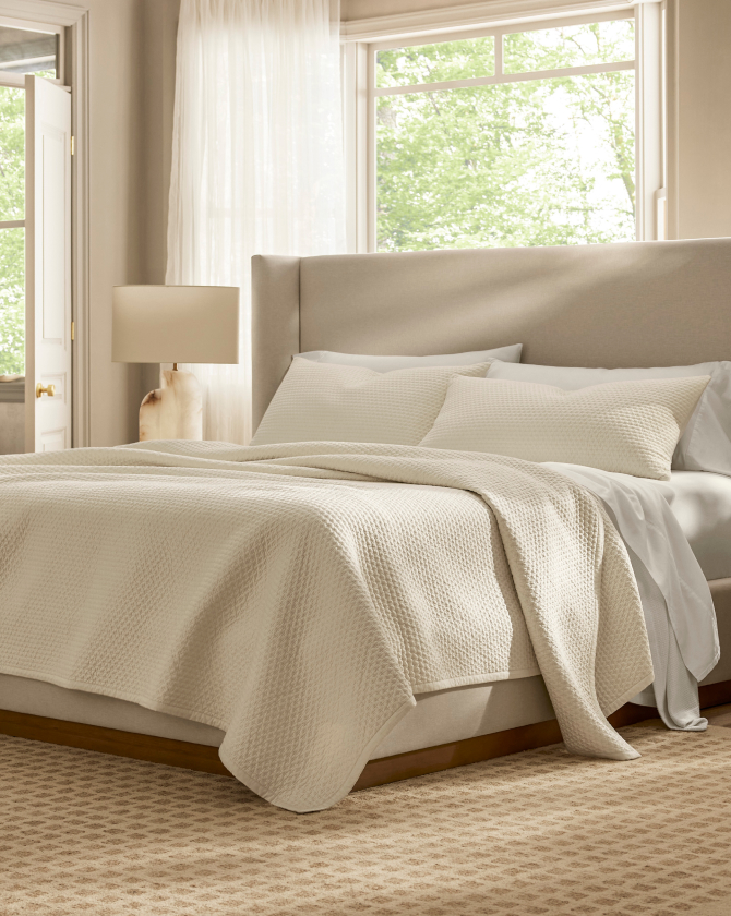 A neatly made bed with white bedding, including a comforter and pillows, is placed in a bright, airy bedroom. A beige upholstered headboard extends the width of the bed. A nightstand with a lamp and a window with sheer curtains and greenery outside are visible.