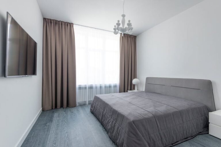 A minimalist bedroom features a queen-sized bed with a grey comforter, a wall-mounted flat-screen TV, and modern light fixtures. A large window with brown curtains partially drawn lets in natural light, while white walls and dark wood floors create a clean look.