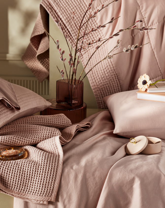 A bedroom scene with a bed covered in soft pink bedding, featuring textured blankets and pillows. A dark glass vase with pussy willow branches sits on a bedside table, alongside books, a flower, and a ring box on the bed, creating a cozy and elegant atmosphere.