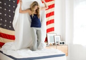 A woman with long blonde hair stands on a bed, holding up a white sheet above her head. She is wearing a navy tank top and gray pajama pants. Behind her is an American flag hung on the wall, and on a dresser nearby are framed pictures and a clock.