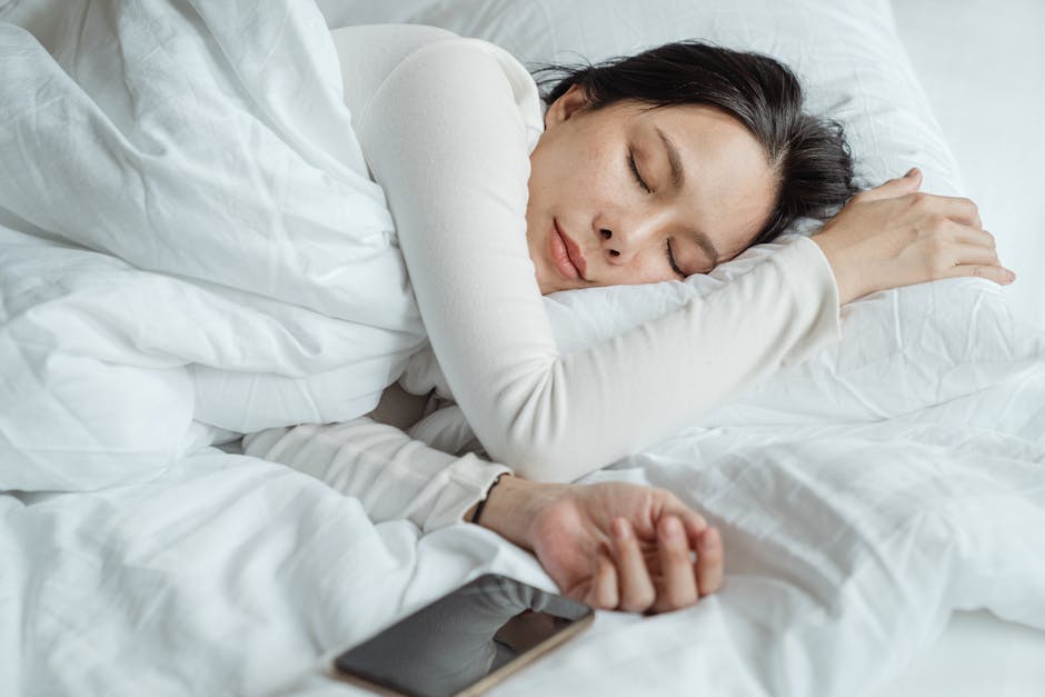 A person peacefully sleeping in a white bed, with their head resting on a pillow and their arm extended near a smartphone lying on the bed. They are wearing a light-colored long sleeve top, and the scene is calm and serene.