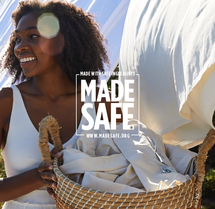 A person with curly hair smiles while holding a basket of laundry. A white sheet billows in the wind in the background. The image features text promoting "MADE SAFE" and safe ingredients, with the website www.madesafe.org.