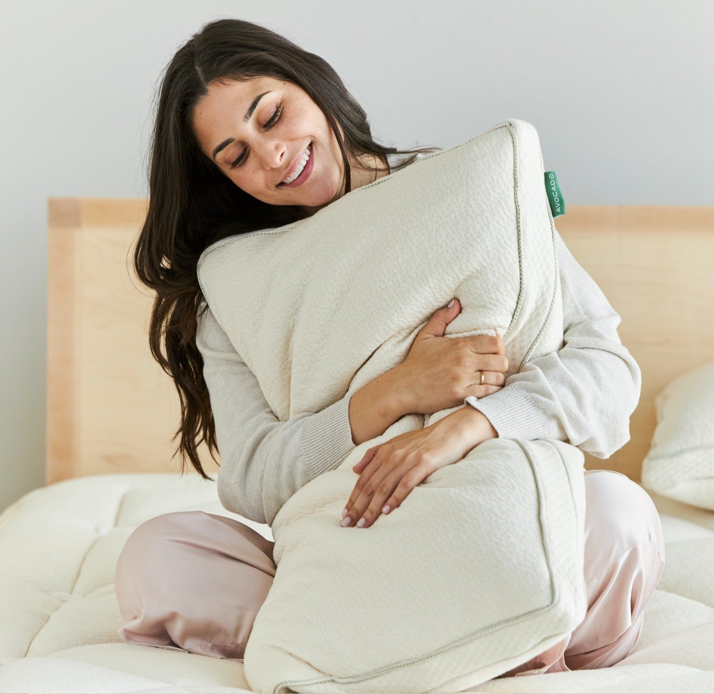 A person with long dark hair is sitting cross-legged on a bed and hugging a large white pillow. They are smiling with their eyes closed, and wearing a light-colored long-sleeve top and light pink pants. The background includes a wooden headboard and a pale wall.