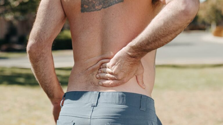 A shirtless person standing outdoors, seen from behind, with their right hand placed on their lower back, appearing to be in discomfort or pain. The person has a tattoo on their upper back and is wearing grayish-blue pants.